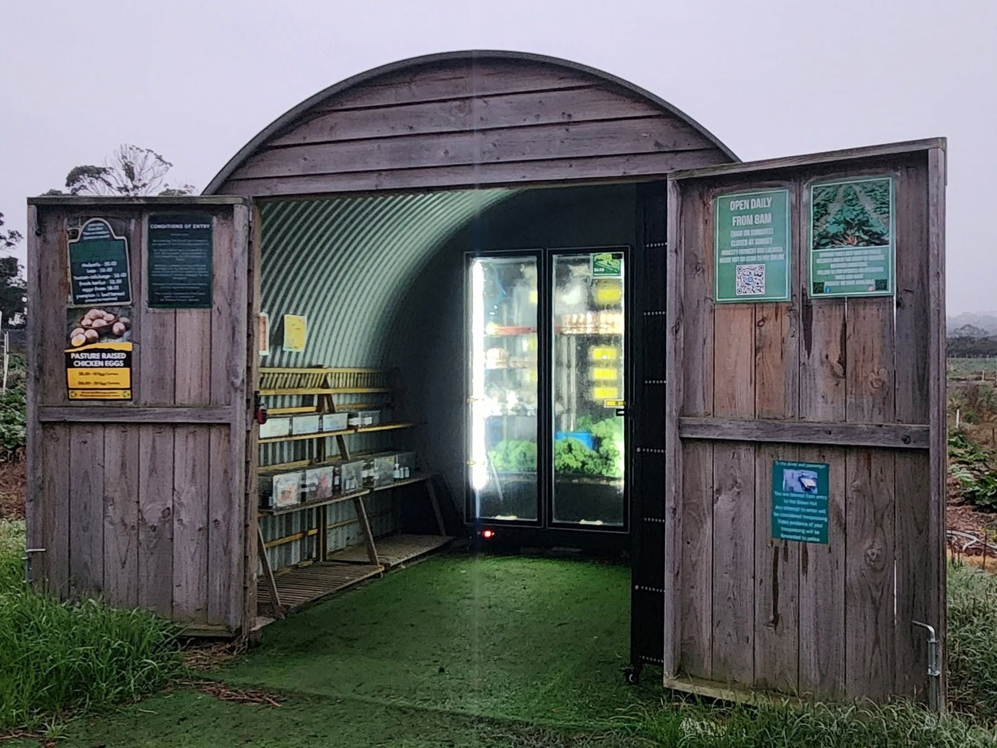 The Green Hut farm gate store — arched corrugated iron hut with open barn doors and lit refrigerators inside
